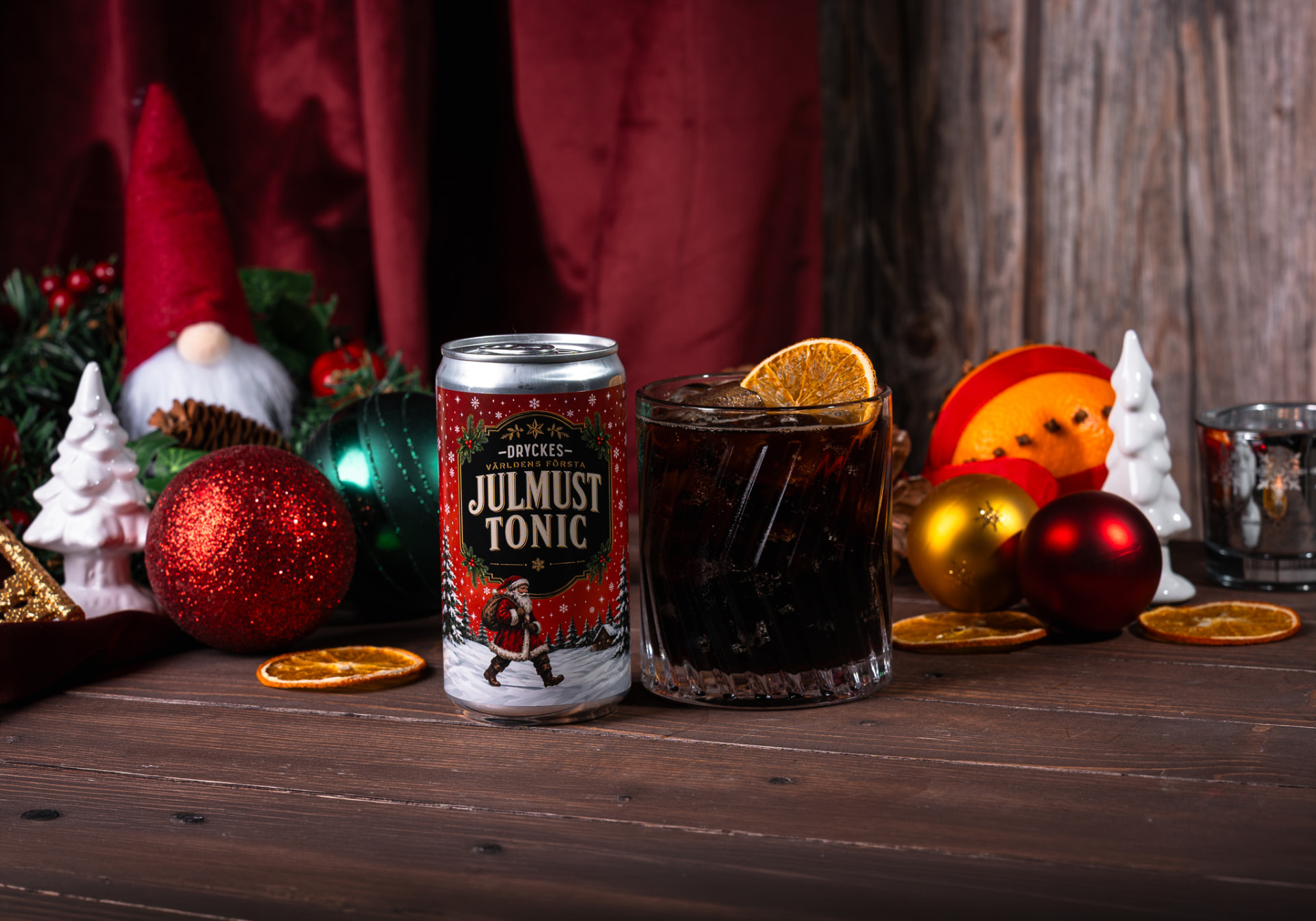 DRYCKES Julmust Tonic-burken bredvid ett mörkt drinkglas med is och apelsinskiva, omgiven av julkulor, torkade apelsinskivor och vinterpynt