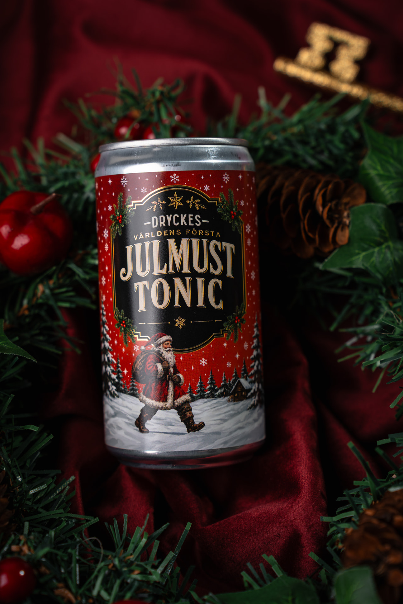 DRYCKES Julmust Tonic-burken framför en julkrans med granris, röda bär, tallkottar och guldkulor mot djupröd sammet